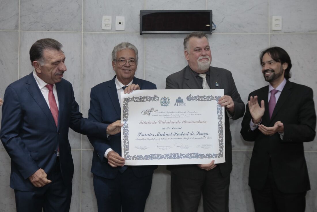 Ranier de barba grisalha segura o diploma. Do lado direito dele o autor da homenagem, deputado Antônio Moraes. Na ponta esquerda deputado eleito José  Patriota e na ponta direita o deputado Diogo Moraes