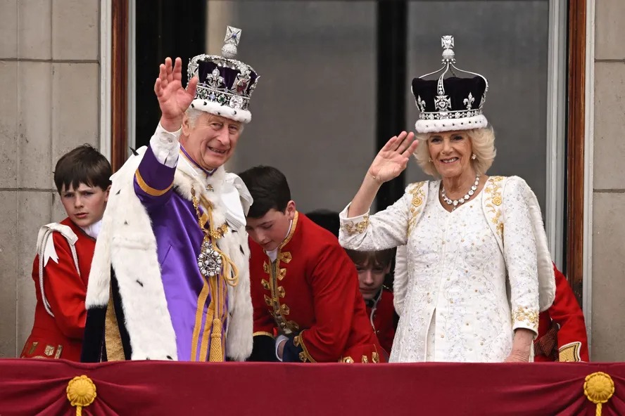 Charles III, vestindo a Coroa do Estado Imperial, e a rainha Camilla, com uma versão modificada da Coroa da Rainha Maria, acenam para os súdito da varanda do Palácio de Buckingham 