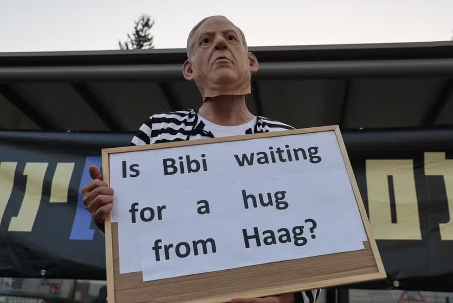 Ativista de esquerda usa máscara do premier de Israel, Benjamin Netanyahu, e roupa de presidiário, e ergue cartaz com dizeres: "Bibi está esperando por um abraço de Haia?"