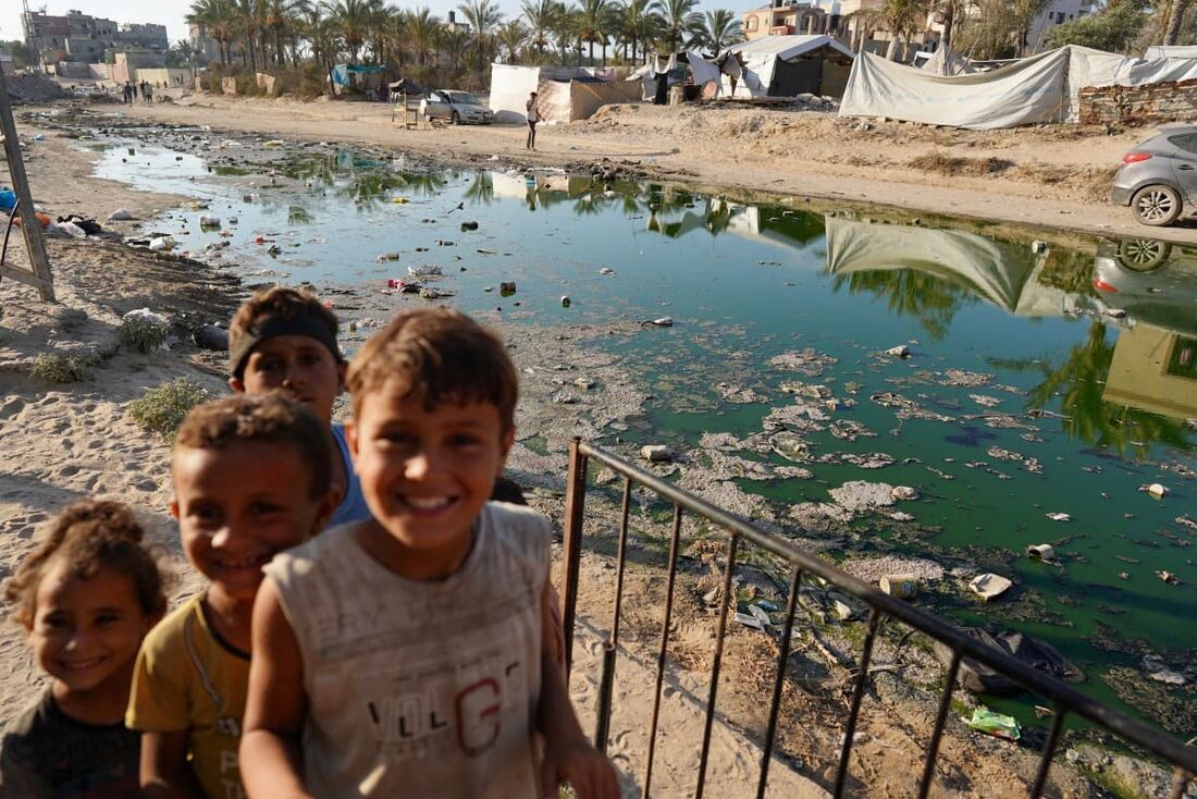  Crianças palestinas sorriem perto de uma rua coberta com águas residuais estagnadas em Deir el-Balah, no centro da Faixa de Gaza