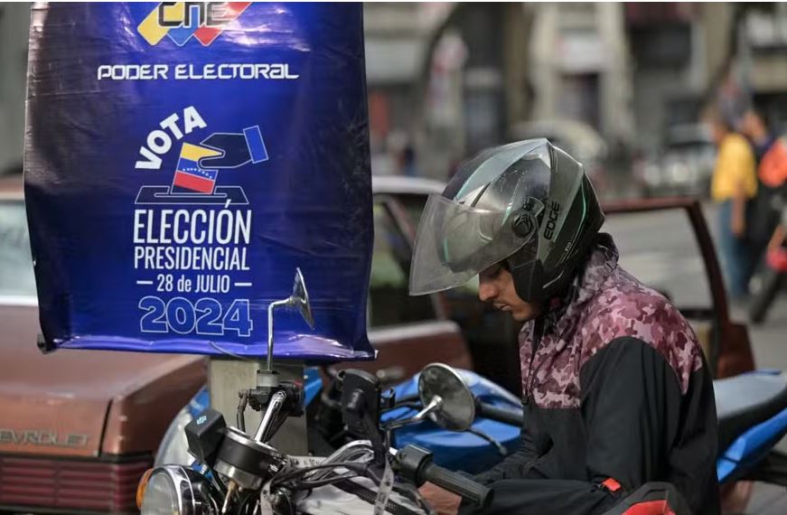 Um motociclista olha seu celular em frente a um cartaz do Conselho Nacional Eleitoral (CNE) promovendo as próximas eleições presidenciais em Caracas, em 23 de julho de 2024 