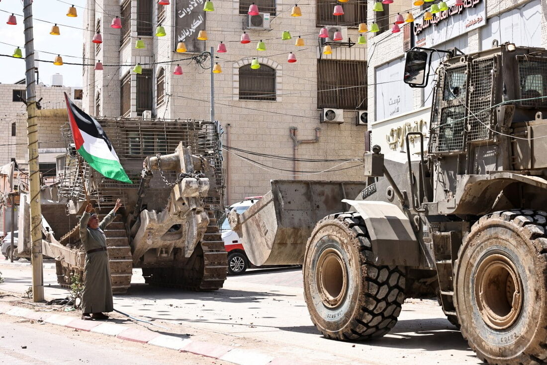 Um homem palestino agita a bandeira da Palestina enquanto veículos do exército israelense bloqueiam a entrada do campo de refugiados palestinos de Tulkarem, na Cisjordânia ocupada