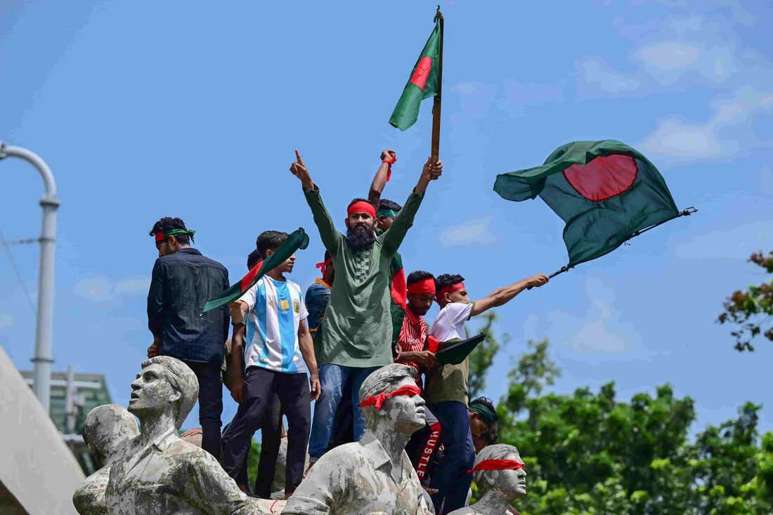 Manifestantes agitam bandeiras nacionais enquanto se posicionam sobre a escultura Memorial Antiterrorismo Raju durante um protesto em Dhaka