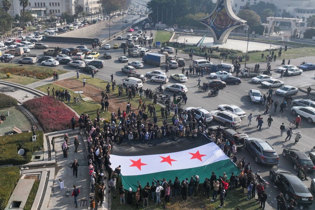 As pessoas comemoram segurando uma grande bandeira da oposição síria na Praça dos Omíadas, em Damasco