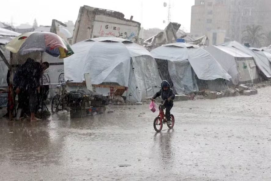 Criança palestina anda de bicicleta passando por tendas encharcadas durante uma tempestade de chuva em um acampamento improvisado na Cidade de Gaza em 31 de dezembro de 2024 