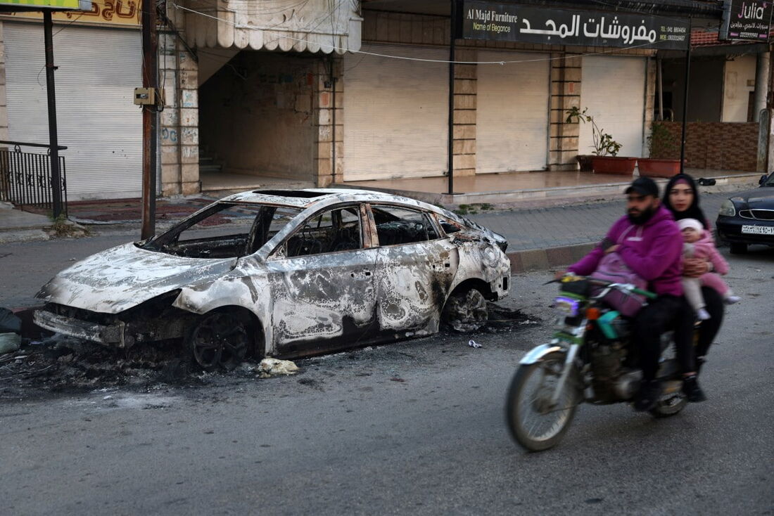 Membros de uma família em uma motocicleta passam por um carro carbonizado na cidade de Jableh, na província costeira de Latakia, na Síria