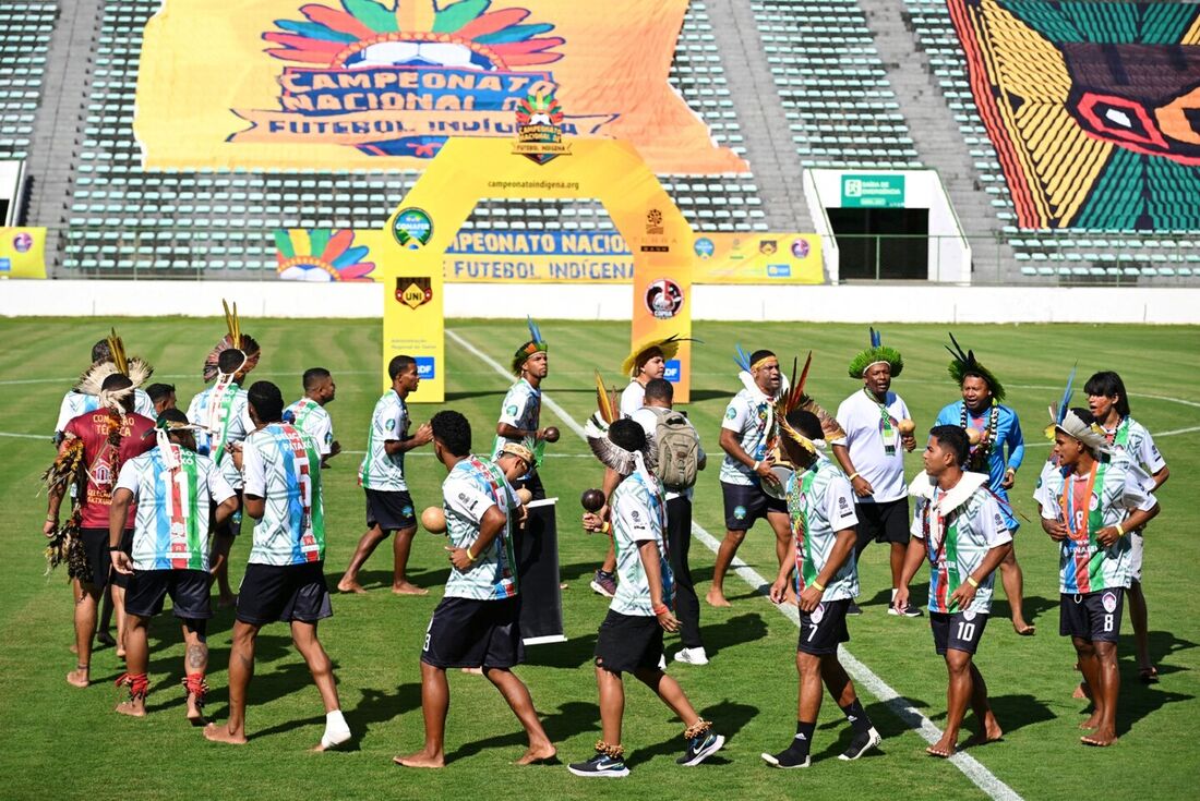 Jogadores com cocares, colares e rostos pintados dobram os joelhos em uma coreografia em um estádio em Brasília