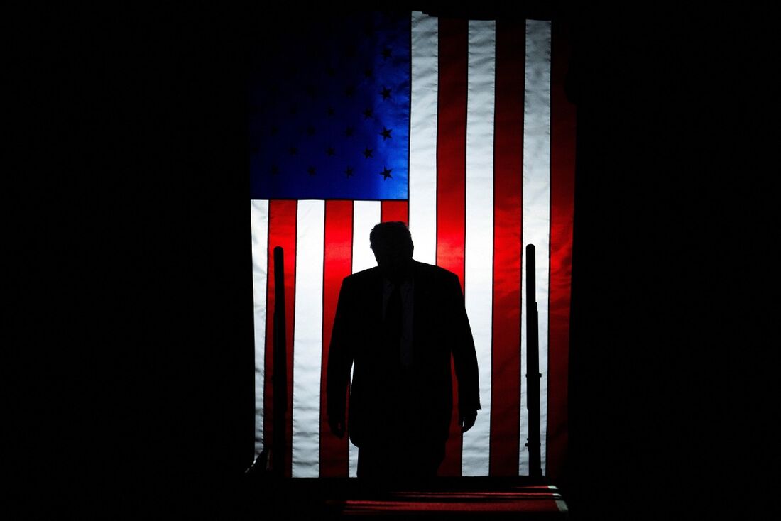 Donald Trump anda na direção da bandeira dos EUA em meio à escuridão