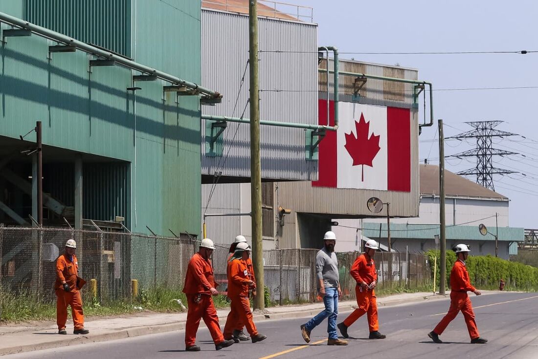 Uma bandeira nacional canadense é vista ao fundo enquanto trabalhadores atravessam a rua em frente aos prédios de fabricação de aço da ArcelorMittal Dofasco em Hamilton, Ontário.