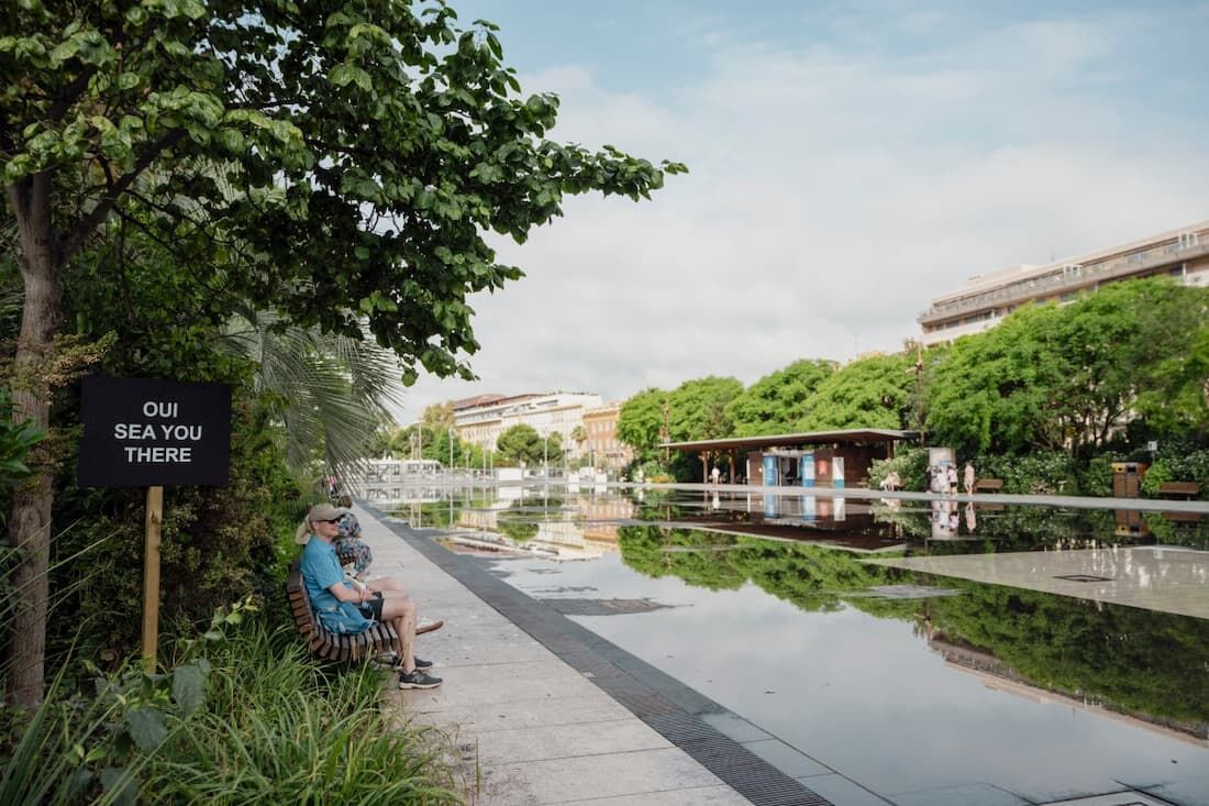 Pessoas sentam-se em um banco perto de uma placa com os dizeres "sim, você está aí" em um parque da cidade em Nice.