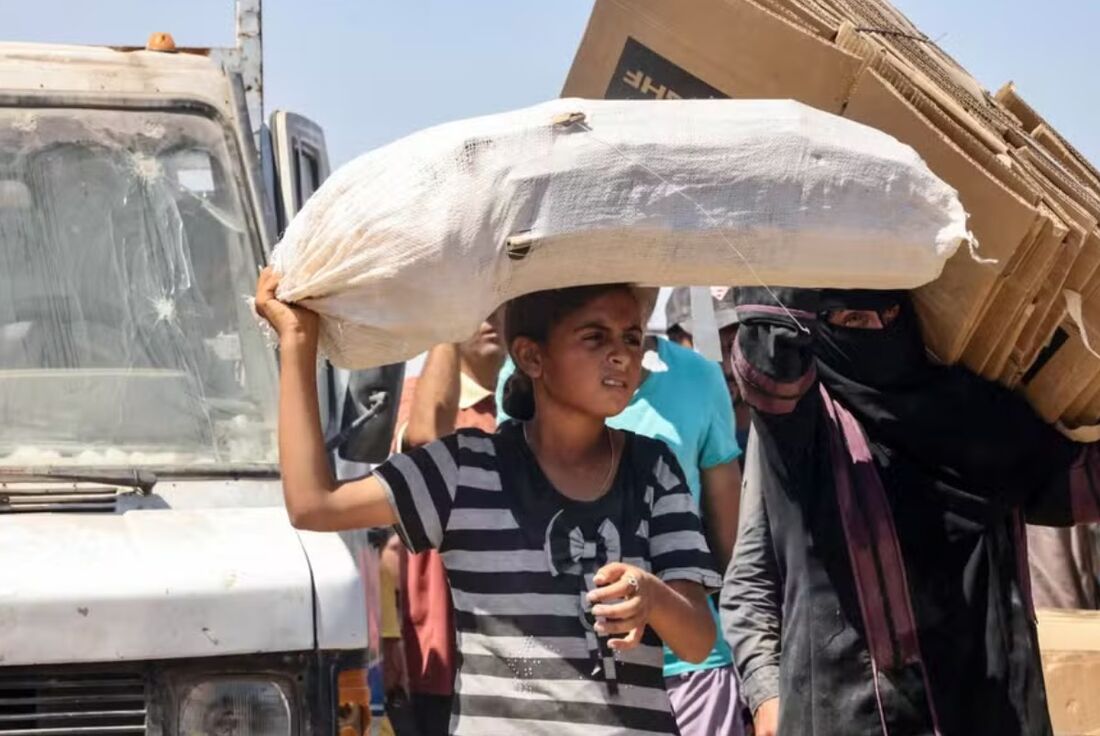 Menina palestina carrega uma sacola enquanto mulher leva caixas vazias no campo de al-Mawasi, em Rafah, no sul de Gaza, em 27 de julho de 2025 