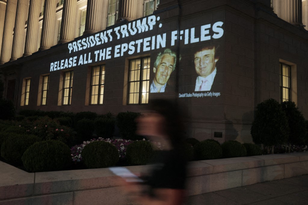 Uma pessoa passa por uma imagem do presidente dos EUA, Donald Trump, e de Jeffrey Epstein, juntamente com as palavras "Presidente Trump: Liberem Todos os Arquivos de Epstein", projetada no prédio da Câmara de Comércio dos EUA
