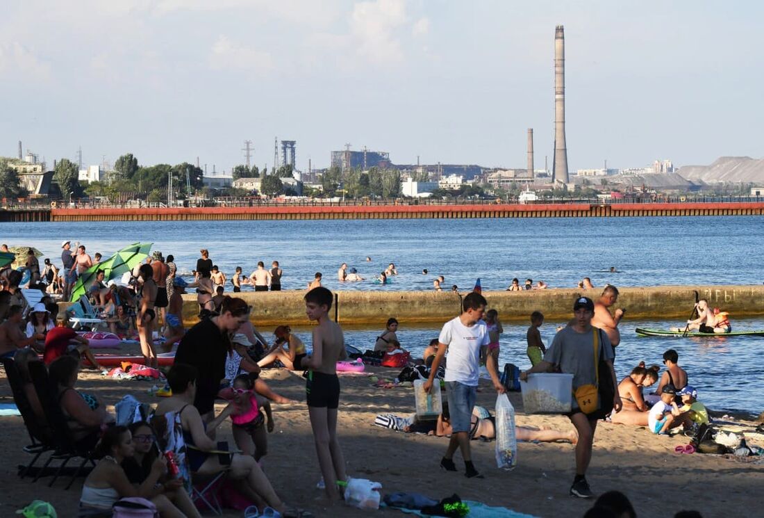 Pessoas aproveitam uma praia na cidade portuária de Mariupol, no Mar de Azov, controlada pela Rússia, no sudeste da Ucrânia, com a usina siderúrgica Azovstal destruída vista à distância