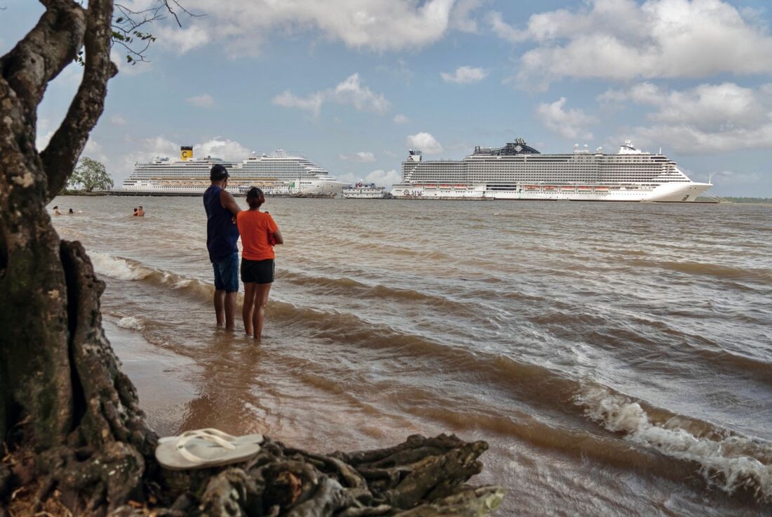 Pessoas em uma praia às margens do Rio Guama observam navios de cruzeiro atracados no Porto de Outeiro, que receberá delegações que participarão da COP30