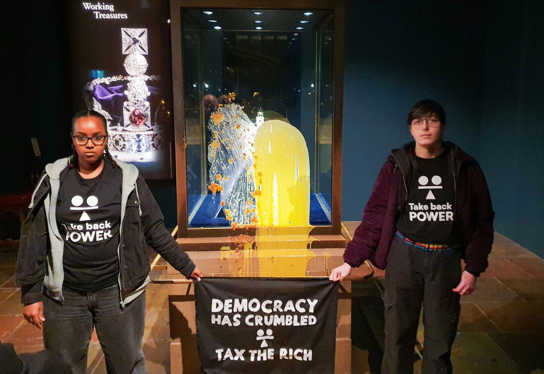 Apoiadores do grupo de resistência civil segurando uma faixa com os dizeres "A democracia desmoronou - Taxem os ricos" após terem jogado sobremesa e creme inglês sobre a vitrine que continha as Joias da Coroa, na Torre de Londres.