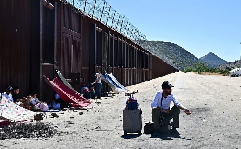 Migrante espera com seus pertences ao lado do muro da fronteira EUA-México após entrar nos EUA vindo do México em Jacumba Hot Springs, Califórnia. Foto: Frederic J. Brown / AFP
