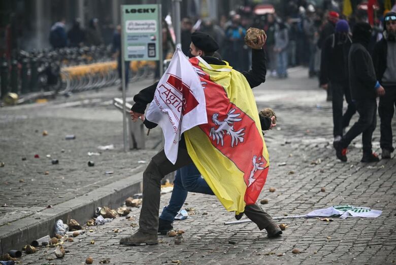 Um manifestante vestindo uma bandeira da Jeunes Agriculteurs (JA, Jovens Agricultores) atira um vegetal perto do Parlamento Europeu.| Foto: AFP